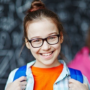 A young girl wearing glasses and a backpack smiling after seeing a children’s dentist in Dallas