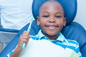 A little boy wearing a striped shirt holding a manual toothbrush and smiling while seated in the dentist’s chair