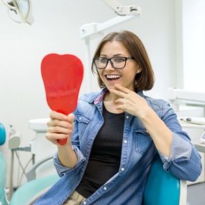 A middle-aged woman wearing glasses looks at her new smile after receiving dental implants in Dallas